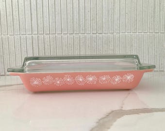 May include: A rectangular, pink Pyrex baking dish with a clear glass lid. The dish is decorated with a white daisy pattern along the side. The dish is approximately 12 inches long.