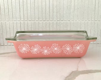 May include: A rectangular, pink Pyrex baking dish with a clear glass lid. The dish features a white daisy pattern along the side. The lid has a green tint. A classic kitchenware item.