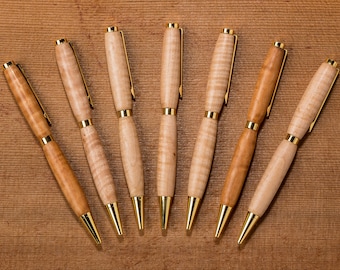 May include: Seven wooden pens with gold accents. The pens are arranged in a semi-circle on a wood surface.