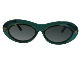 May include: Oval sunglasses with a translucent dark teal plastic frame and dark gray lenses. Each temple hinge area features a silver-toned letter M emblem. The design has a smooth, rounded silhouette with a bridge connecting the two oval lenses.