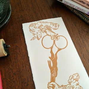 May include: A gold-colored print of a hand holding two rings, with a cloud-like design above and below the hand.