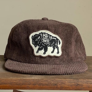 Op de afbeelding: Bruine corduroy baseballcap met een zwart-wit buffelpatch. De cap heeft een gebogen klep en een knoop aan de bovenkant. De hoed is gemaakt van een geribbelde corduroy stof.