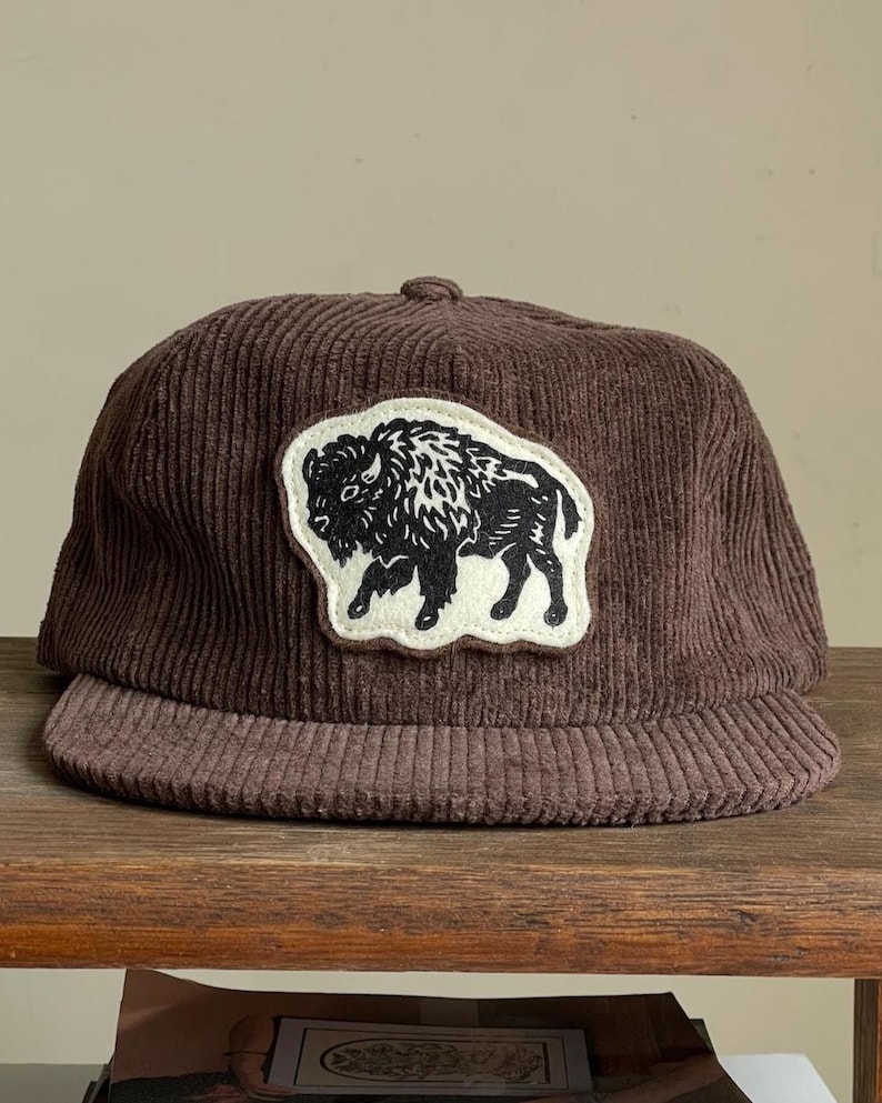 Op de afbeelding: Bruine corduroy baseballcap met een zwart-wit buffelpatch. De cap heeft een gebogen klep en een knoop aan de bovenkant. De hoed is gemaakt van een geribbelde corduroy stof.