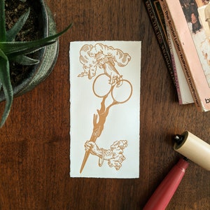 May include: A brown and gold print of a hand holding a pair of scissors with a cloud-like design above the hand.