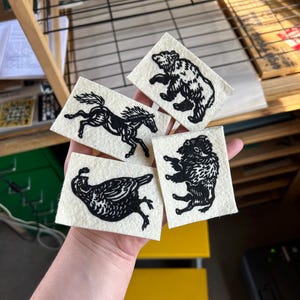 Op de afbeelding: Een hand houdt vier kleine, rechthoekige stoffen patches vast met zwarte linoleumsnede afdrukken van dieren. De afbeeldingen tonen een beer, een paard, een vogel en een bizon. De patches bevinden zich op een lichte achtergrond.