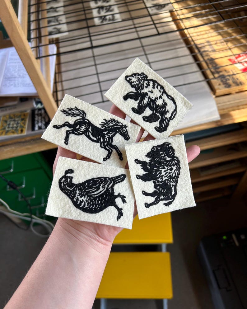 Op de afbeelding: Een hand houdt vier kleine, rechthoekige stoffen patches vast met zwarte linoleumsnede afdrukken van dieren. De afbeeldingen tonen een beer, een paard, een vogel en een bizon. De patches bevinden zich op een lichte achtergrond.