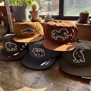 Peut inclure: Une collection de cinq casquettes de baseball dans divers tons terreux, notamment moutarde, marron, olive et gris. Chaque casquette présente un écusson brodé unique sur le panneau avant représentant un poisson, un cheval, un bison, un ours ou une caille.