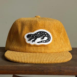 Op de afbeelding: Mosterdgele corduroy baseballpet met een zwart-witte hagedis patch. De pet heeft een gebogen klep en een gestructureerde kroon. De hagedis patch is gecentreerd op de voorkant.