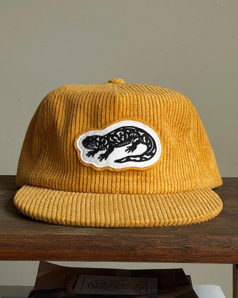 Op de afbeelding: Mosterdgele corduroy baseballpet met een zwart-witte hagedis patch. De pet heeft een gebogen klep en een gestructureerde kroon. De hagedis patch is gecentreerd op de voorkant.