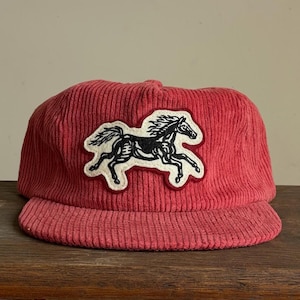 Op de afbeelding: Een rode corduroy baseballcap met een zwart-wit paardenpatch. De cap heeft een gebogen klep en een gestructureerde kroon. De paardenpatch is een gedetailleerde illustratie van een rennend paard.