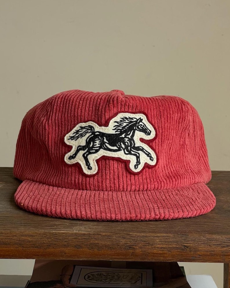 Op de afbeelding: Een rode corduroy baseballcap met een zwart-wit paardenpatch. De cap heeft een gebogen klep en een gestructureerde kroon. De paardenpatch is een gedetailleerde illustratie van een rennend paard.