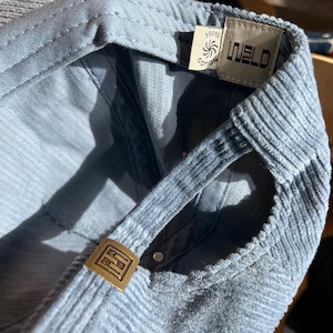 Op de afbeelding: Close-up van een lichtblauwe corduroy baseballpet. De pet heeft een gebogen klep, verstelbare band en een metalen gesp. De binnenkant toont een label met de tekst "WELD" en "100% Cotton".