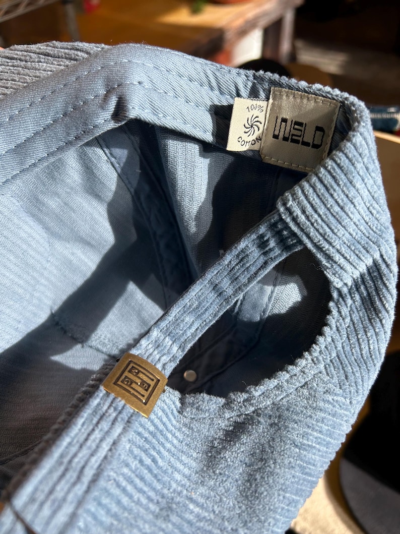 Op de afbeelding: Close-up van een lichtblauwe corduroy baseballpet. De pet heeft een gebogen klep, verstelbare band en een metalen gesp. De binnenkant toont een label met de tekst "WELD" en "100% Cotton".