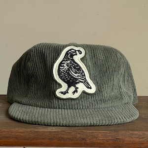 Op de afbeelding: Een saliegroene corduroy baseballcap met een gebogen klep. Een witte patch met een zwarte vogelillustratie is op de voorkant genaaid. De hoed wordt gepresenteerd op een houten oppervlak.