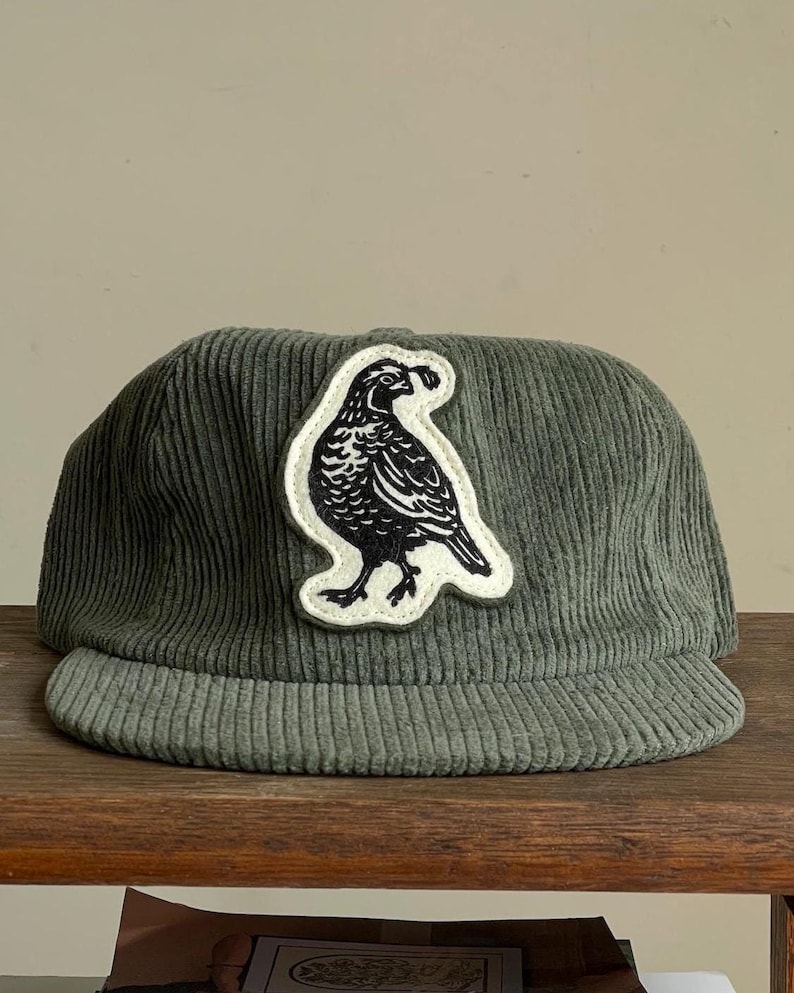 Op de afbeelding: Een saliegroene corduroy baseballcap met een gebogen klep. Een witte patch met een zwarte vogelillustratie is op de voorkant genaaid. De hoed wordt gepresenteerd op een houten oppervlak.