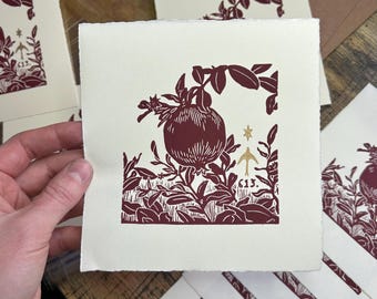 May include: Square cream print with a burgundy botanical design of a pomegranate and foliage. The print includes the number "613" and a gold star. Several similar prints and envelopes are visible.