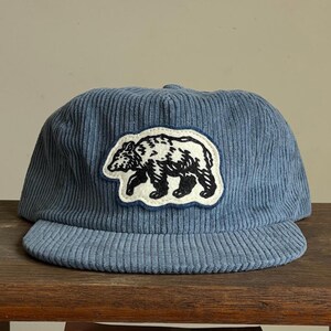Op de afbeelding: Blauwe corduroy baseballcap met een zwart-witte berenpatch. De cap heeft een gebogen klep en een gestructureerde kroon. De berenpatch is wit omlijnd en heeft een gedetailleerde berenillustratie.