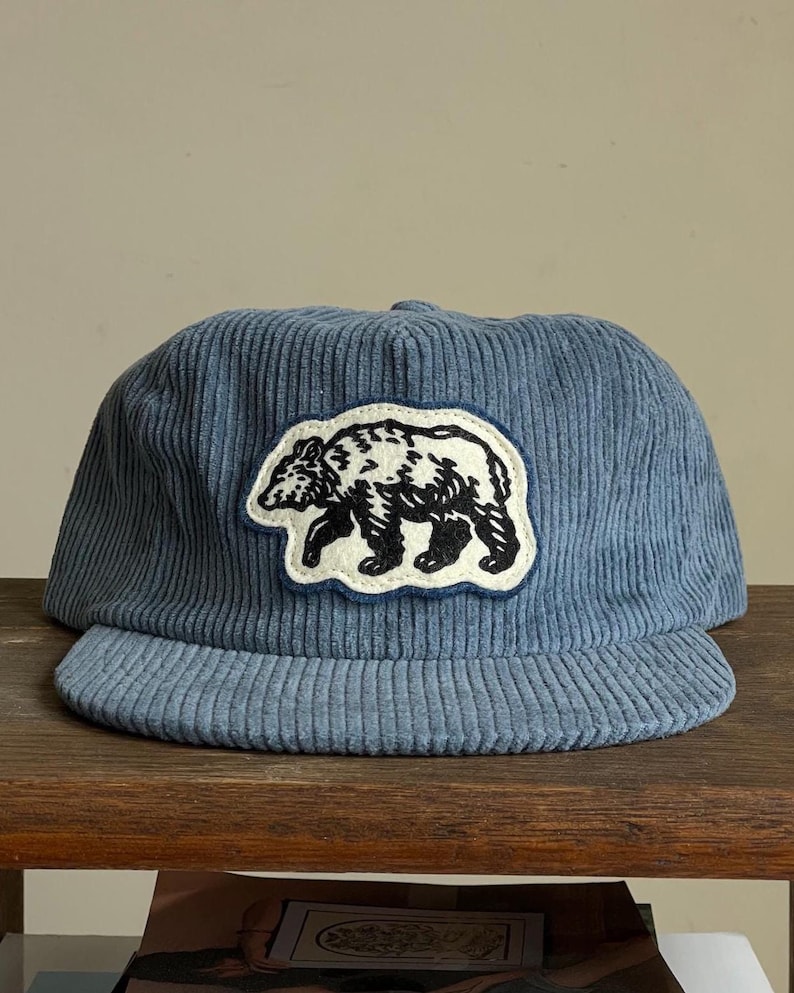Op de afbeelding: Blauwe corduroy baseballcap met een zwart-witte berenpatch. De cap heeft een gebogen klep en een gestructureerde kroon. De berenpatch is wit omlijnd en heeft een gedetailleerde berenillustratie.