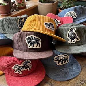 May include: A collection of corduroy baseball caps in various colors, including yellow, red, and blue. Each cap features a patch with a different animal illustration, such as a bear, horse, or bison. The caps have a classic, casual style.