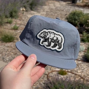 May include: A slate blue baseball cap featuring a white and black embroidered patch of a walking bear on the front panel. The cap is constructed from a lightweight, crinkled fabric and includes a flat brim with detailed stitching along the edge.