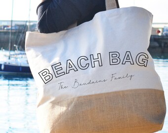 May include: A large, off-white and tan beach bag with the words "BEACH BAG" in bold, black letters. Below the text is the phrase "The Baudains Family" in a smaller, cursive font. The bag has a natural-colored bottom and handles.