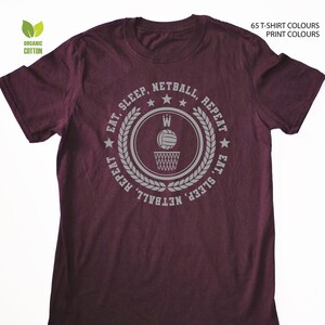 May include: Burgundy t-shirt with a white graphic of a basketball hoop and ball with the text "Eat, Sleep, Netball, Repeat" around the graphic.