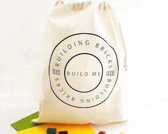 May include: A white canvas drawstring bag with a black circle logo that says "Building Bricks Build Me Building Bricks". The bag is filled with colorful Lego bricks.
