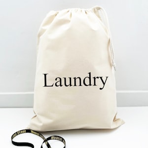 Puede incluir: Una bolsa de lavandería de lona blanca con un cordón de ajuste negro y la palabra "Laundry" impresa en negro en la parte delantera.