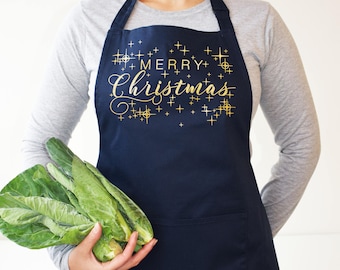 Op de afbeelding: Een donkerblauwe schort met een gouden print die "Merry Christmas" zegt met gouden sterren en plustekens.