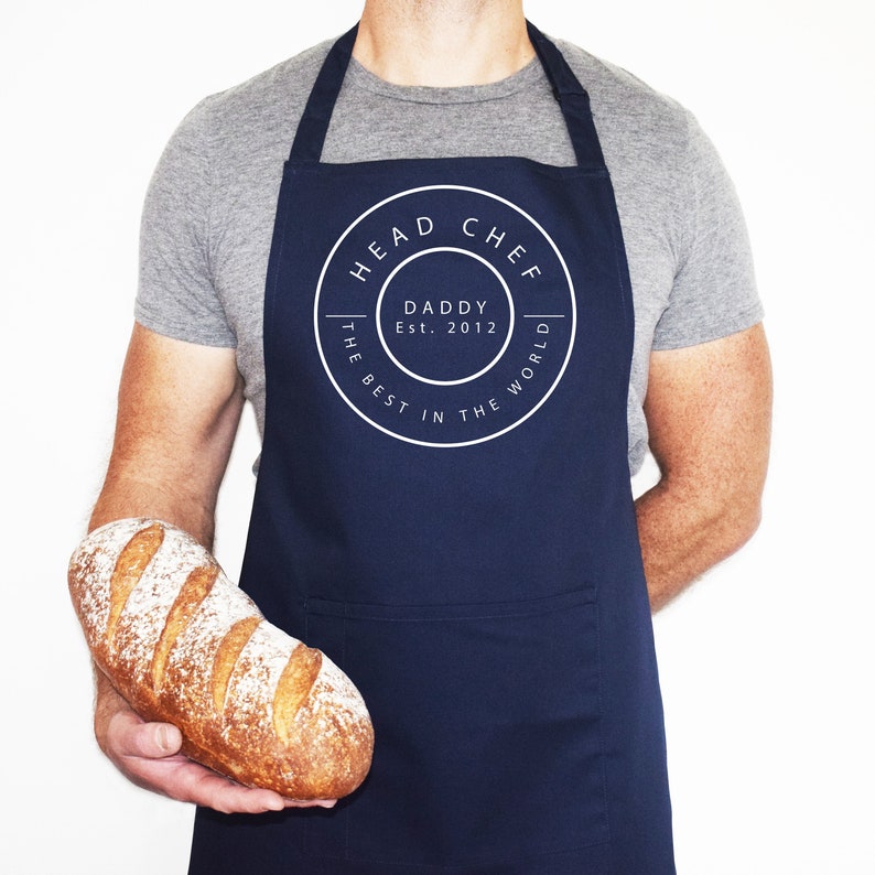 An apron made from polyester with a front pocket, sliding adjustable buckle on the neckband so the wearer can adjust the overall length with custom name, date print on the solid color background