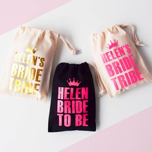 Könnte beinhalten: Drei kleine Stoffbeutel mit Kordelzug. Zwei Beutel sind weiß mit goldener Aufschrift "Helen's Bride Tribe". Ein Beutel ist schwarz mit rosa Aufschrift "Helen Bride To Be". Alle Beutel haben ein kleines Kronen-Motiv.