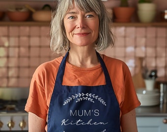 May include: A woman wearing a blue apron with the text "Mum's Kitchen" in white lettering and a wreath of leaves around the text. The apron has a white background and is worn over an orange shirt.