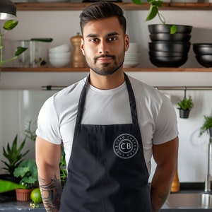 Puede incluir: Un hombre con una camiseta blanca y un delantal negro con el texto "Head Chef" y "CB" en un círculo en la parte delantera.