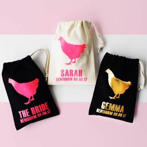 May include: Three black drawstring bags with pink and gold chicken silhouettes. The bags are personalized with the names "Sarah", "Gemma", and "The Bride" and the location "Benidorm 08.08.17".