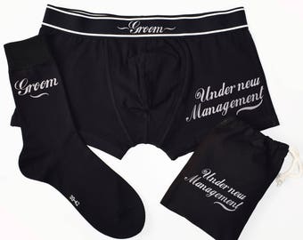May include: Black boxer briefs with a white waistband and the text "Groom" on the waistband. The boxer briefs also have the text "Under new Management" on the front. A black sock with the text "Groom" on it and a black drawstring bag with the text "Under new Management" are also included.