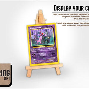 May include: A purple and yellow Pokemon card featuring a pink and black Pokemon with the text "The Anniversary 200 HP". The card is displayed on a wooden easel.