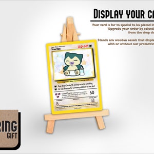 May include: A yellow and white Pokemon card featuring a cartoon Snorlax with a baby pacifier. The card is displayed on a wooden easel stand. The text on the card reads "Snorlax 2024 HP".