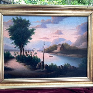 May include: A landscape painting in a gold-toned frame depicting a river scene at sunset. The composition features a small house on a grassy bank, a lone boat on the water, a large tree in the foreground, and distant mountains under a pink sky.