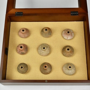 Puede incluir: Una caja de madera con tapa de vidrio muestra nueve peque&ntilde;os objetos redondos de cer&aacute;mica. Los objetos est&aacute;n decorados con varios patrones y dise&ntilde;os en tonos marrones, blancos y rojos.