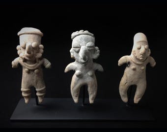 Pode incluir: Três pequenas figuras de aspeto antigo, com formas humanas estilizadas. As figuras são bege claro, com traços faciais e detalhes corporais simples. Estão sobre uma base preta, sobre um fundo escuro.
