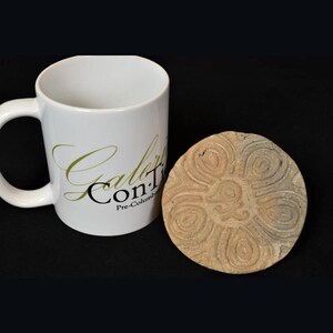 May include: A round, brown, ceramic stamp with a carved design of a flower with six petals. The stamp is sitting on a black surface next to a white coffee mug with the text "Galeria Con T" in green and black.
