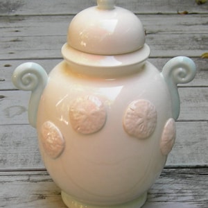 May include: White ceramic jar with a lid and blue handles. The jar is decorated with raised sand dollar designs.