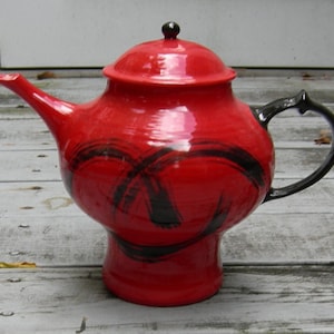 May include: A red teapot with a black handle and black brushstrokes on the side. The teapot has a round body and a tall spout.