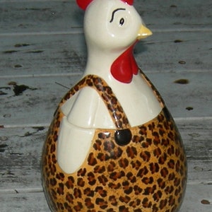 Puede incluir: Una figurita de pollo de cer&aacute;mica blanca con cresta y barbilla rojas, que lleva un mono de leopardo marr&oacute;n y negro.