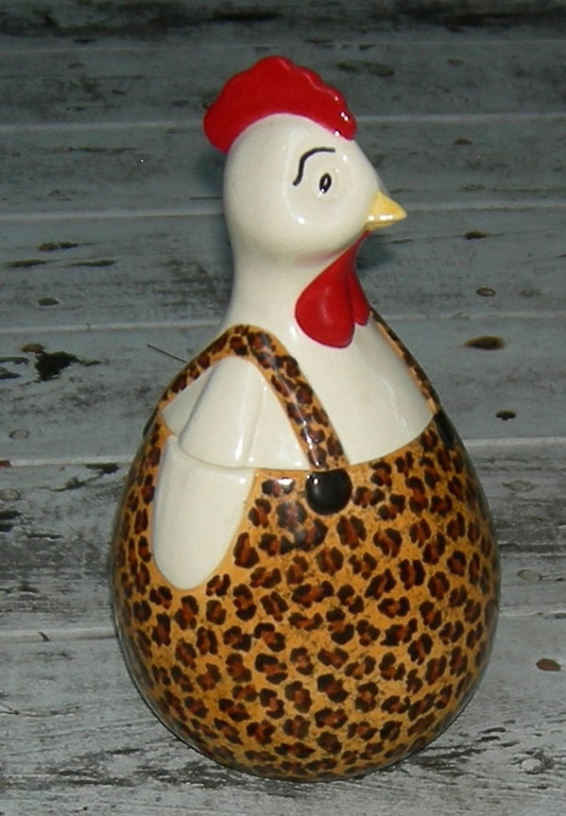 Puede incluir: Una figurita de pollo de cer&aacute;mica blanca con cresta y barbilla rojas, que lleva un mono de leopardo marr&oacute;n y negro.