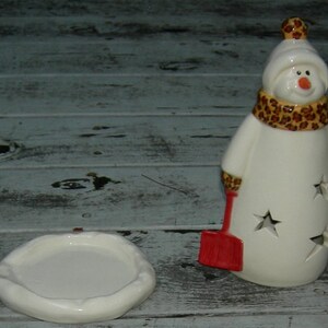 Può includere: Portacandele in ceramica bianca a forma di pupazzo di neve con una sciarpa a motivi marroni e neri e una pala rossa. Il pupazzo di neve ha un design a forma di stella ritagliata ed è seduto su una base in ceramica bianca.