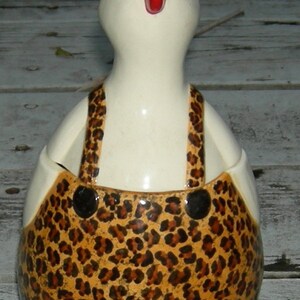 Puede incluir: Una figurita de pollo de cer&aacute;mica blanca con una cresta roja y un patr&oacute;n de leopardo marr&oacute;n y negro en su cuerpo. El pollo lleva un mono.
