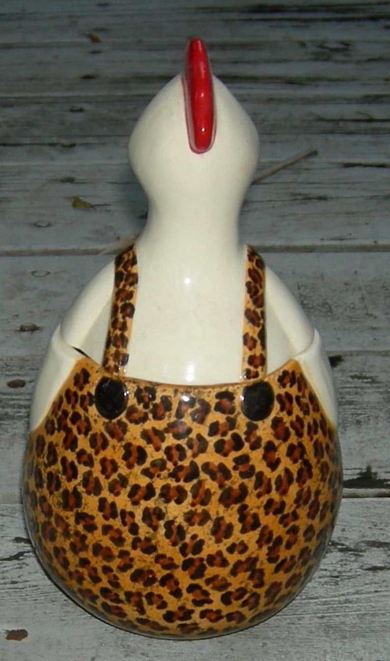 Puede incluir: Una figurita de pollo de cer&aacute;mica blanca con una cresta roja y un patr&oacute;n de leopardo marr&oacute;n y negro en su cuerpo. El pollo lleva un mono.