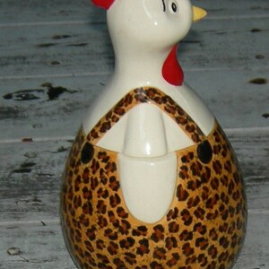Puede incluir: Una figurita de pollo de cer&aacute;mica con una cresta roja y un dise&ntilde;o de estampado de leopardo. El pollo est&aacute; de pie sobre una superficie de madera blanca.
