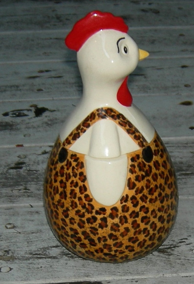 Puede incluir: Una figurita de pollo de cer&aacute;mica con una cresta roja y un dise&ntilde;o de estampado de leopardo. El pollo est&aacute; de pie sobre una superficie de madera blanca.